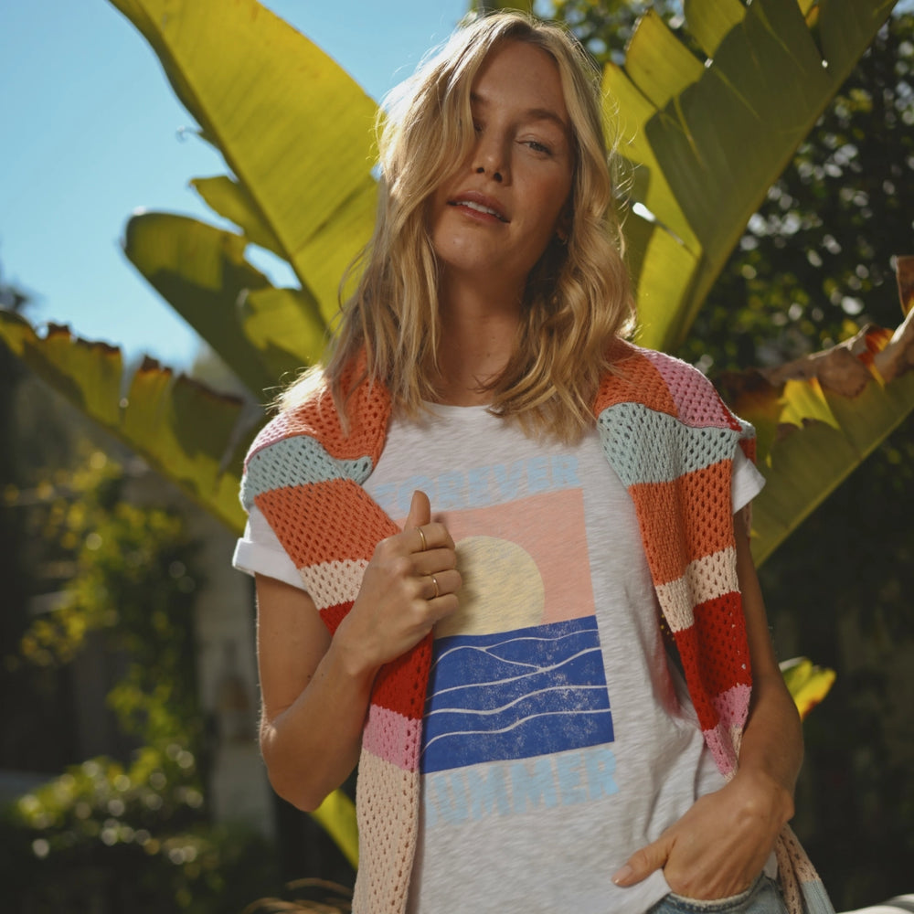 A young woman with wavy blonde hair stands in front of a lush background with large green tropical leaves. She’s wearing a white t-shirt with a colorful graphic of a sunset over ocean waves and the word “SUMMER,” paired with a multi-colored crocheted vest draped over her shoulders. Her expression is relaxed, and she holds the edge of her vest with one hand. The overlaid text reads “SHOP NEW.”