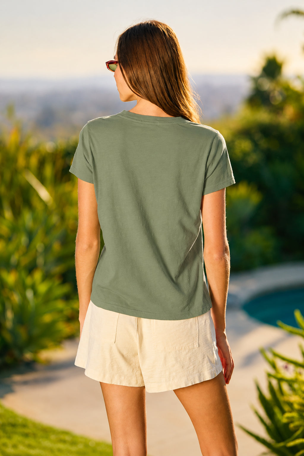 A woman viewed from behind wearing a bright lime green short-sleeve t-shirt and off-white shorts, standing outdoors near a pool with a blurred garden background.