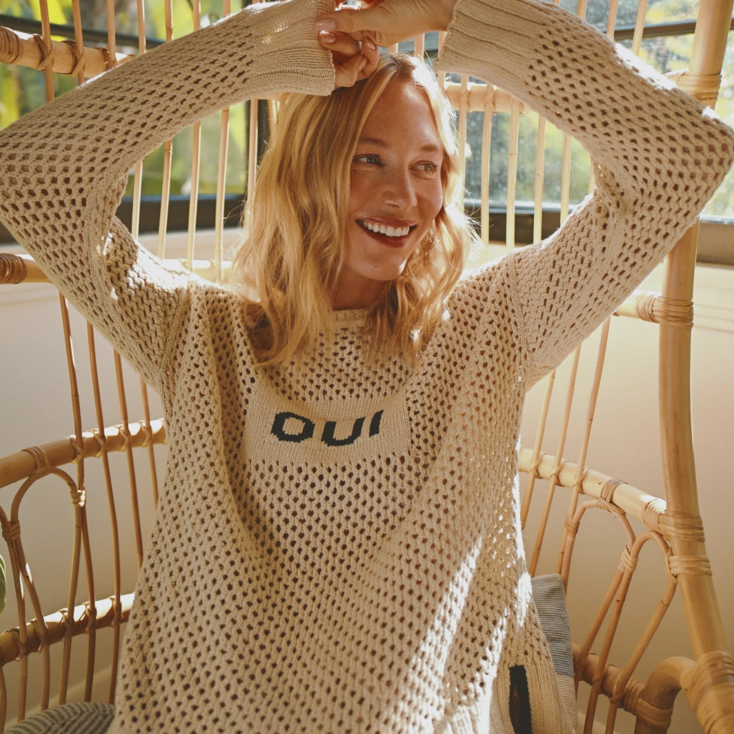 A young woman with wavy blond hair sits in a wicker chair with her arms raised above her head, smiling joyfully. She is wearing a loose, open-knit cream-colored sweater over a top that says "oui." The background features warm, natural light and a wooden-framed window with leafy greenery outside. The overlaid text reads “SHOP SWEATERS.”
