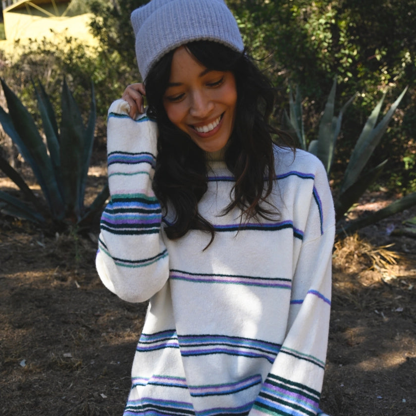 Woman wearing a white sweater with blue and green stripes and a gray beanie, standing outdoors.
