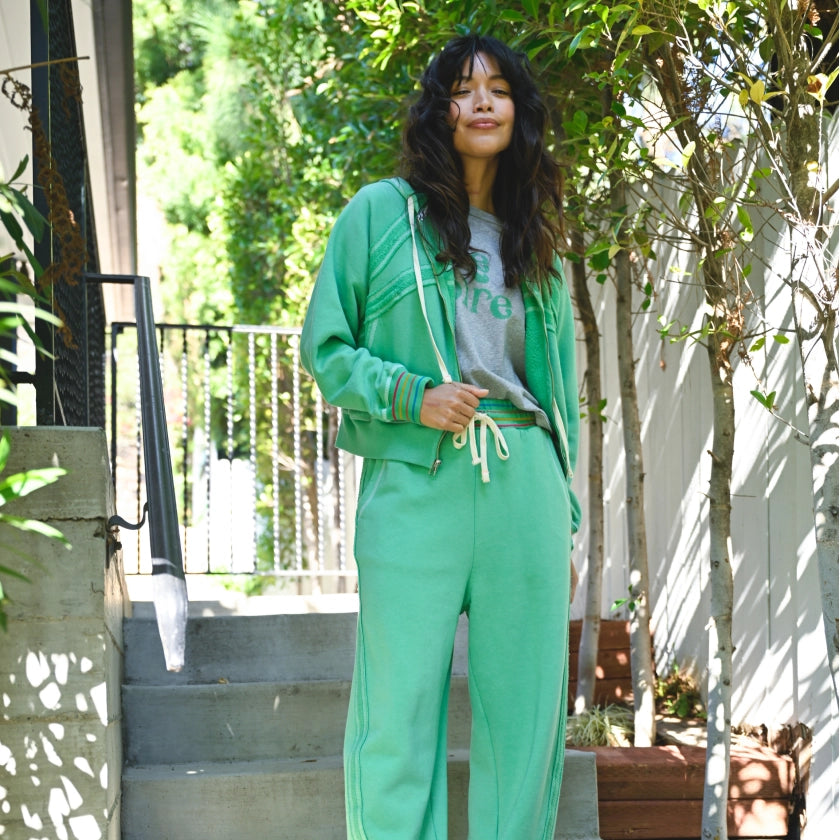 Woman in a green tracksuit standing outdoors with trees and a white wall in the background