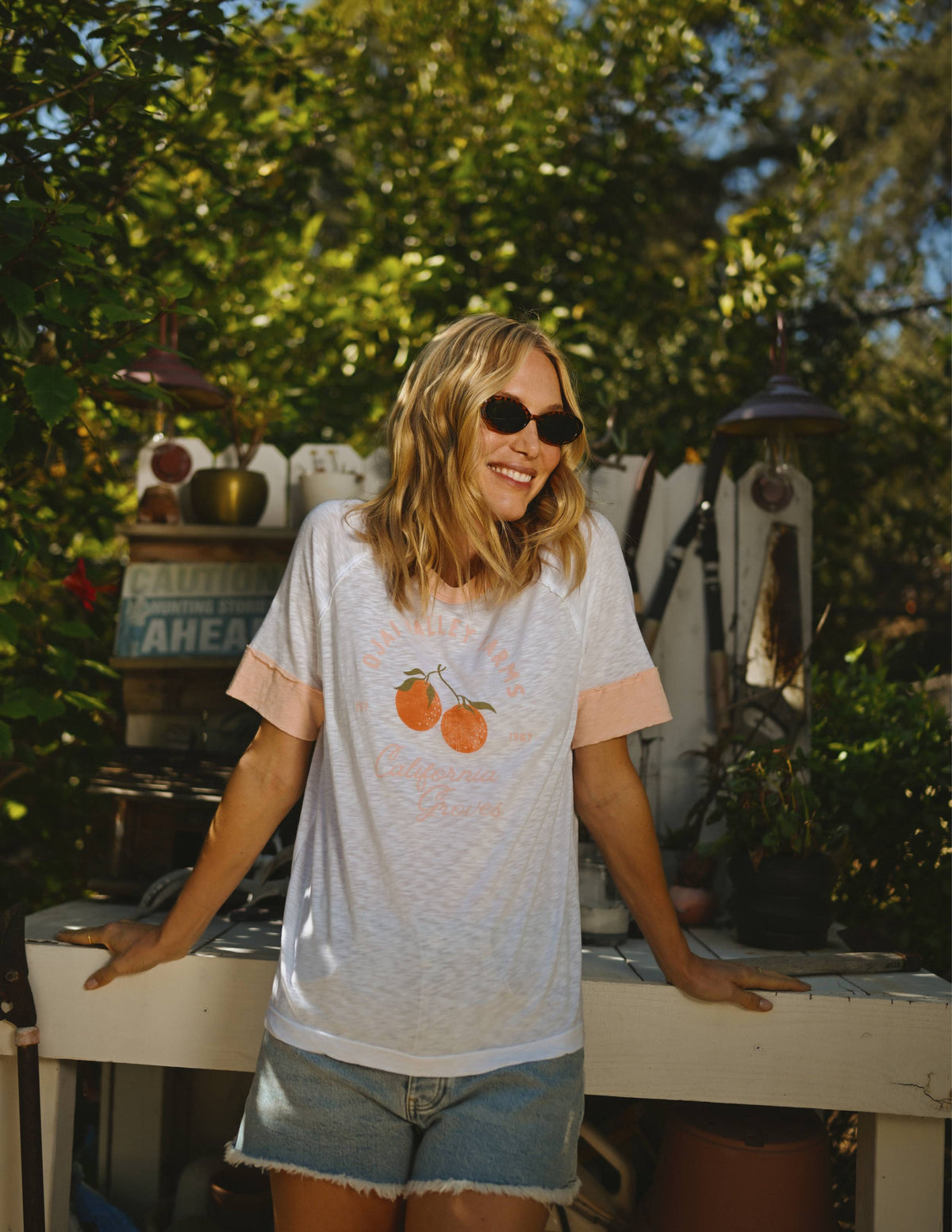 A woman smiling and wearing a white t-shirt with a graphic, blue denim shorts, sunglasses, and standing in front of a garden backdrop.