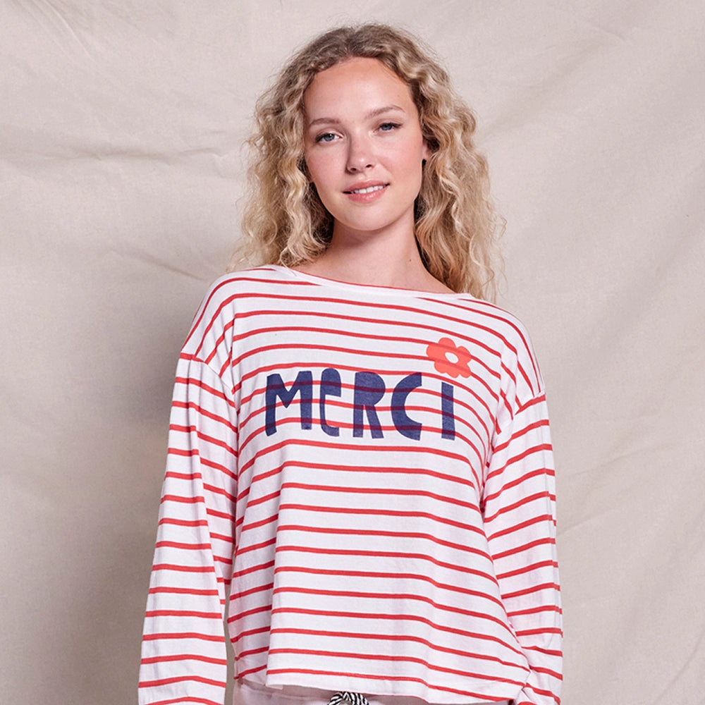 Woman wearing a red and white striped long-sleeve top with the word 'MERCI' in bold navy letters, standing against a neutral fabric background.