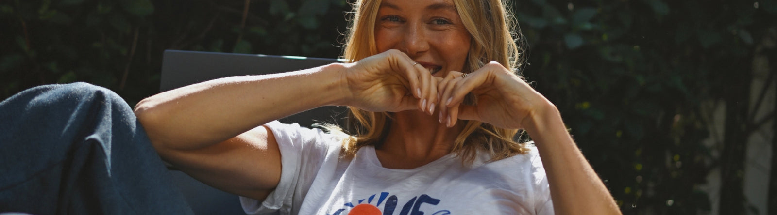  A young woman with wavy blond hair sits outdoors, leaning back in a relaxed position. She wears a white t-shirt with a colorful graphic design and has her arms playfully bent in front of her mouth, smiling warmly. The leafy green foliage in the background is softly blurred, and overlaid text reads “NEW ARRIVALS.”