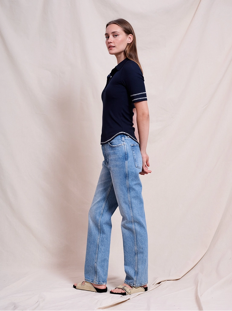 Woman wearing a navy polo shirt with striped sleeve accents and light blue jeans, standing sideways against a beige fabric backdrop