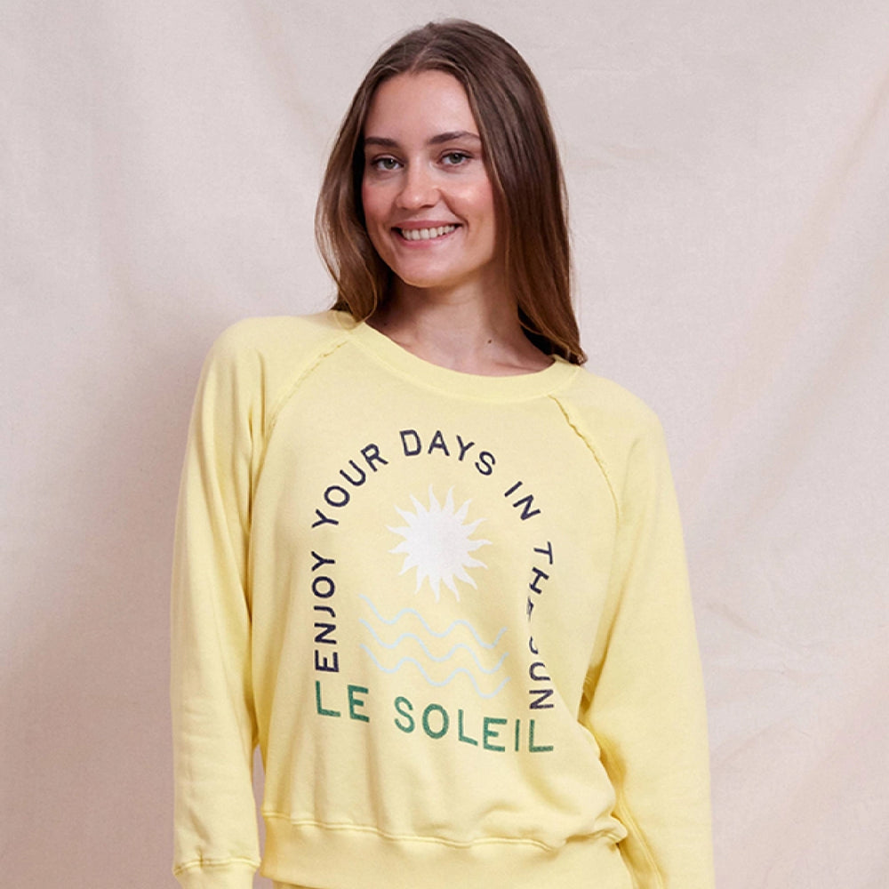 Woman wearing a light yellow sweatshirt with 'ENJOY YOUR DAYS IN THE SUN - LE SOLEIL' graphic, smiling in front of a neutral fabric backdrop.