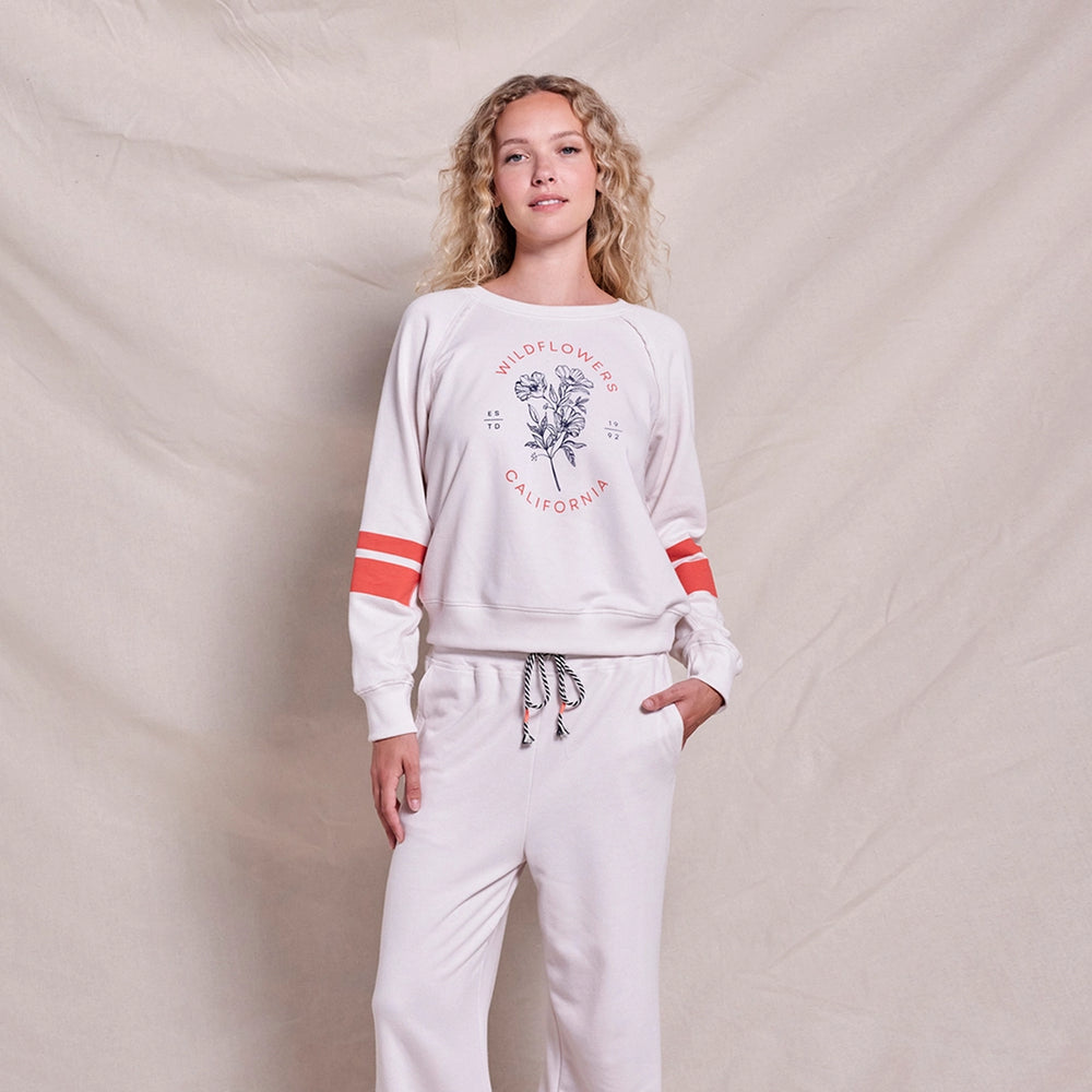 Woman wearing a light-colored lounge set with red-striped sleeves and a 'Wildflowers California' graphic on the sweatshirt, standing against a neutral fabric backdrop.