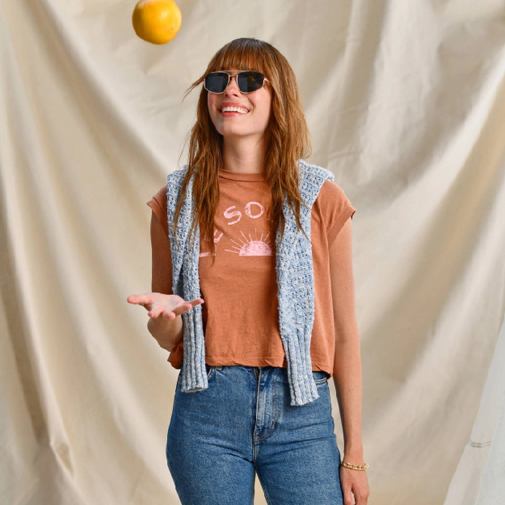Smiling woman in sunglasses wearing a brown cropped tee with 'SO' text, blue jeans, and a sweater draped over her shoulders, tossing an orange against a beige fabric backdrop.